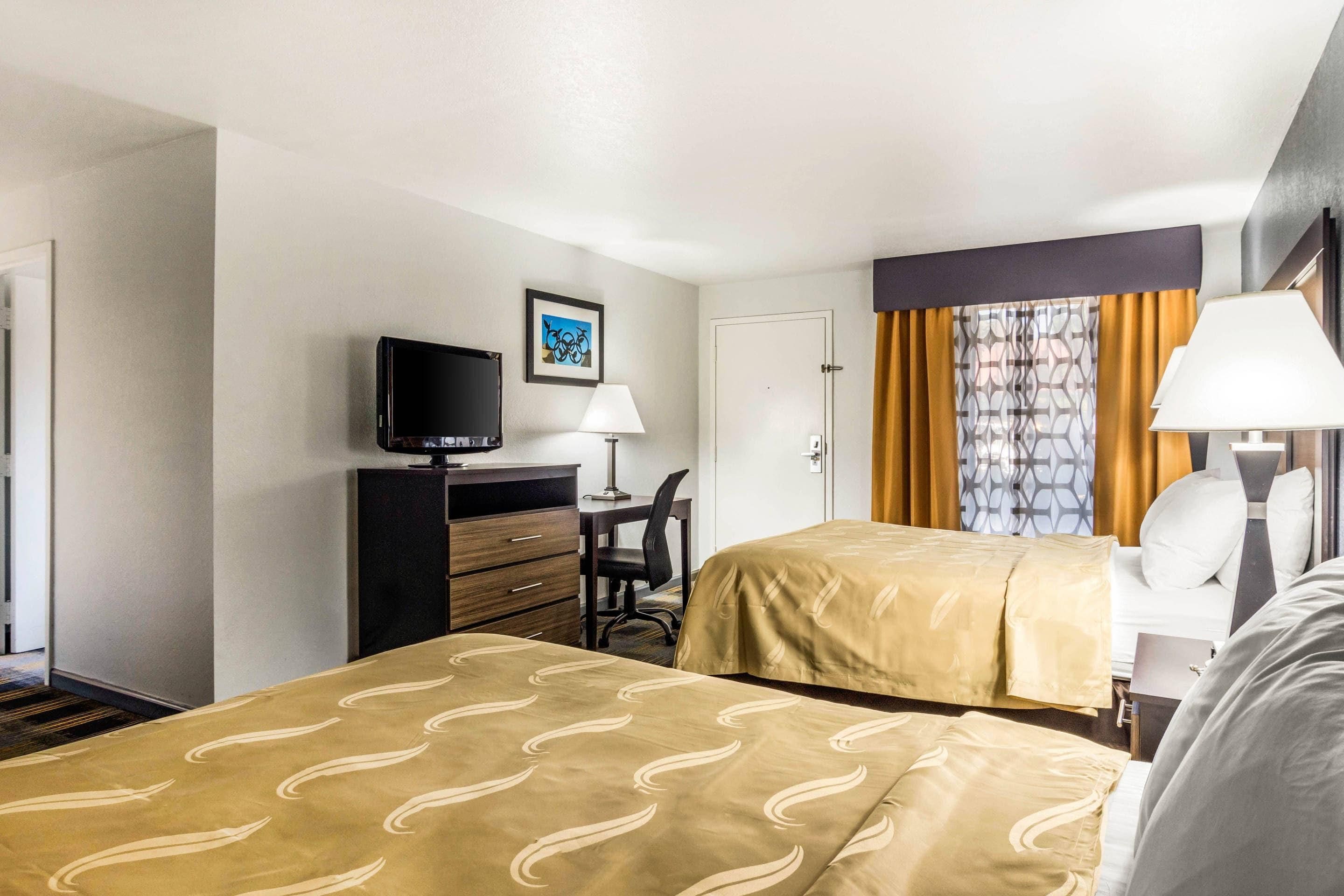 Ein Hotelzimmer mit zwei Betten, einem Schreibtisch mit Computer, einem Fernseher und einem Fenster mit Vorhängen.