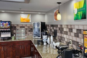 Um saguão de hotel com máquina de café, bebedouro e área de estar com cadeiras e mesa.