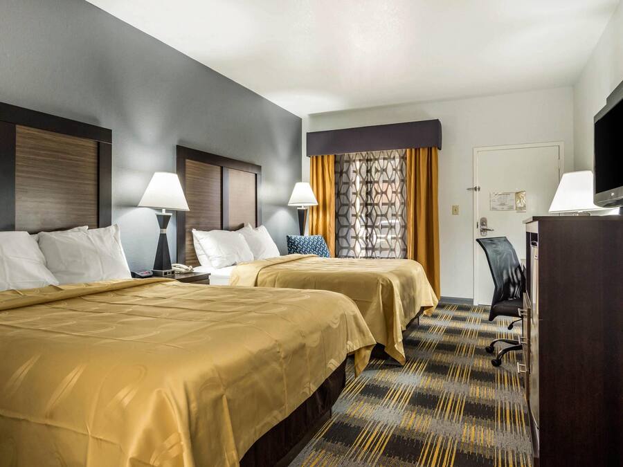 Quarto de hotel com duas camas, uma escrivaninha, uma cadeira, uma televisão e uma porta.