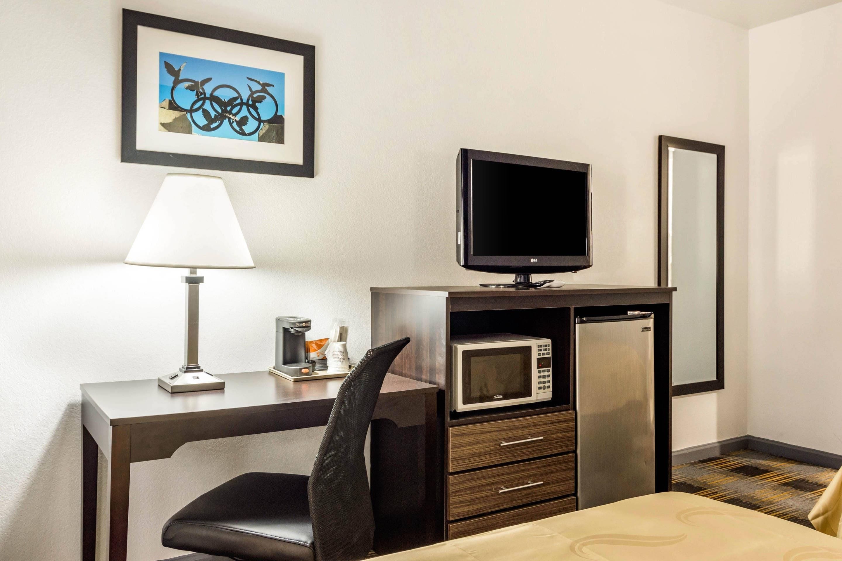 Habitación de hotel con escritorio, silla, televisor, microondas, refrigerador, mesita de noche y cama.