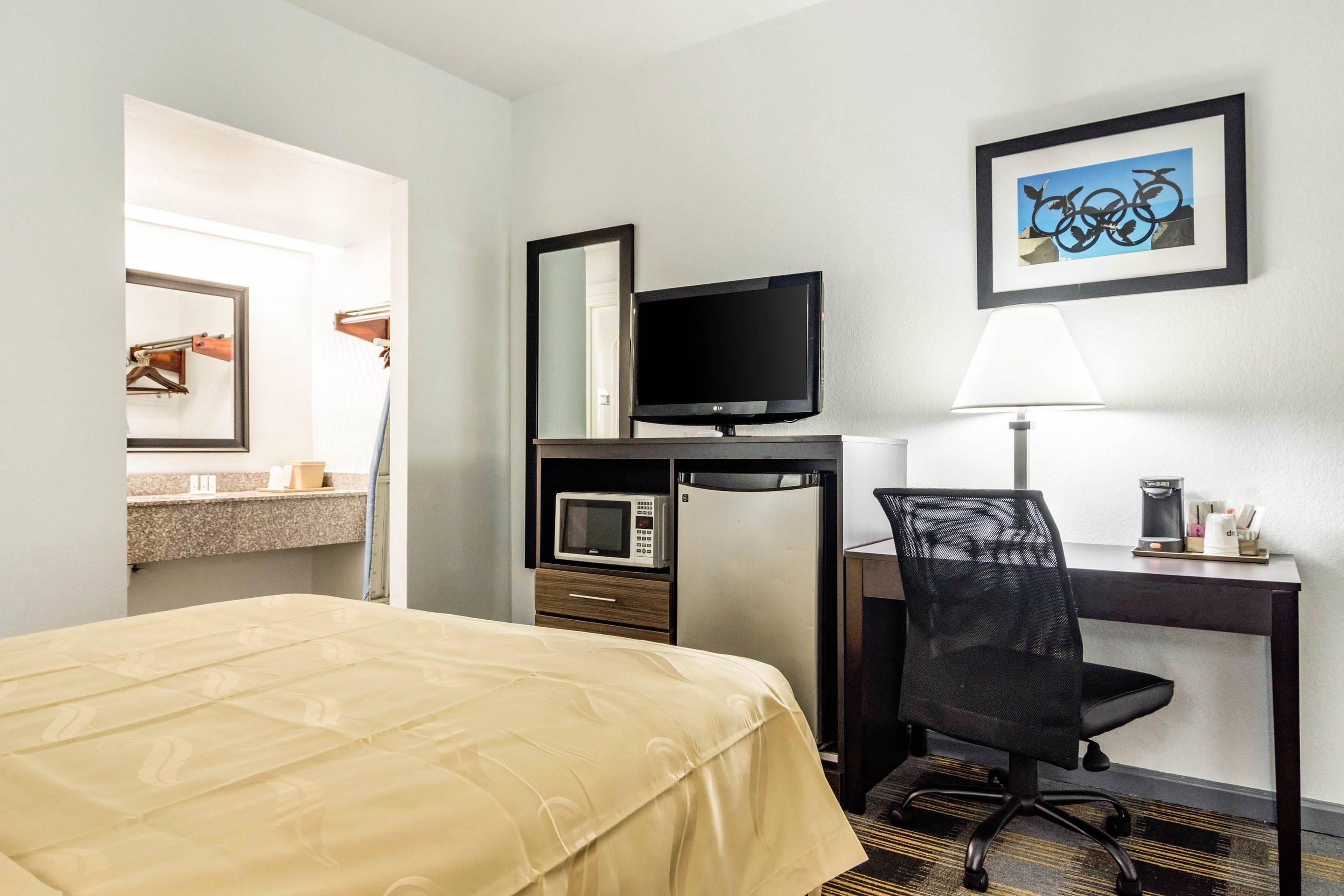Habitación de hotel con cama, escritorio con computadora, televisor, microondas, silla y un cuadro en la pared.