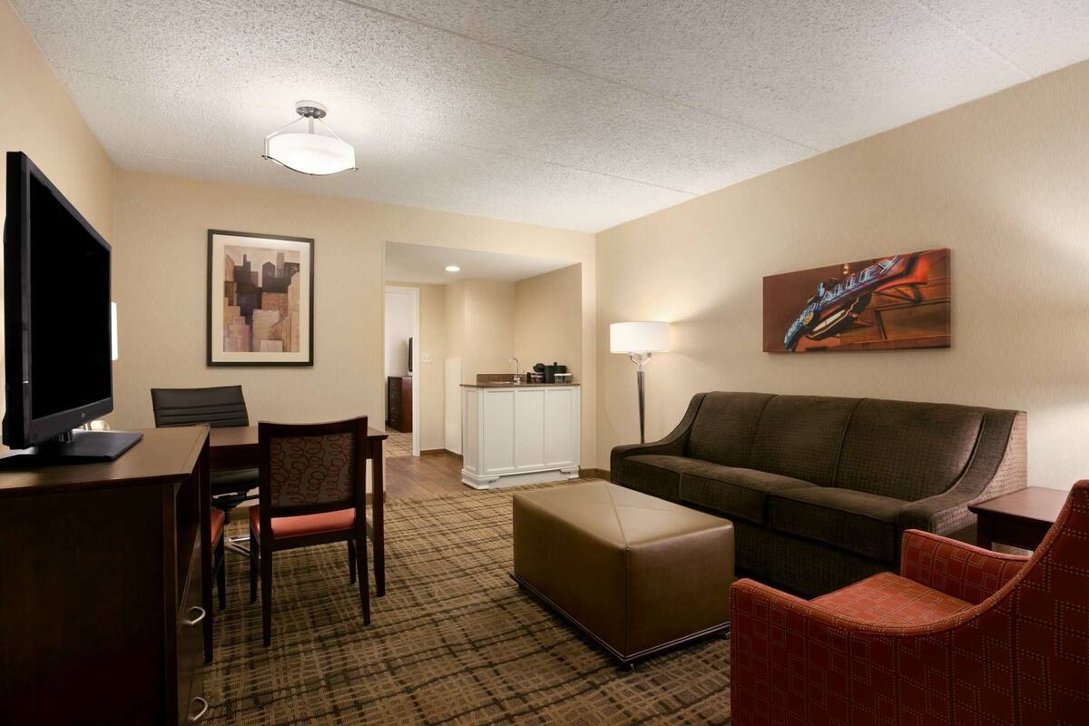 Habitación de hotel con televisión de pantalla plana, escritorio con silla, sofá, otoman y lámpara, además de obras de arte en las paredes.