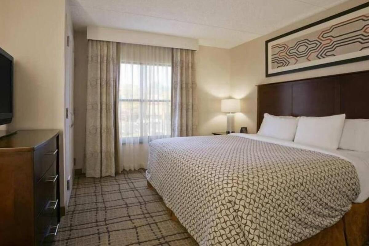 Una habitación de hotel con una cama, televisor, escritorio y ventana con cortinas.