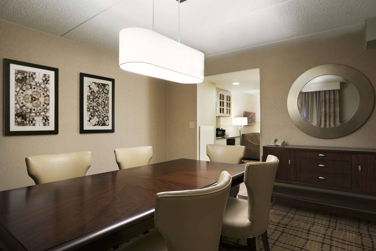 Un comedor de hotel con una gran mesa de madera, sillas color beige, obras de arte enmarcadas en las paredes, un espejo redondo y un aparador de madera oscura.