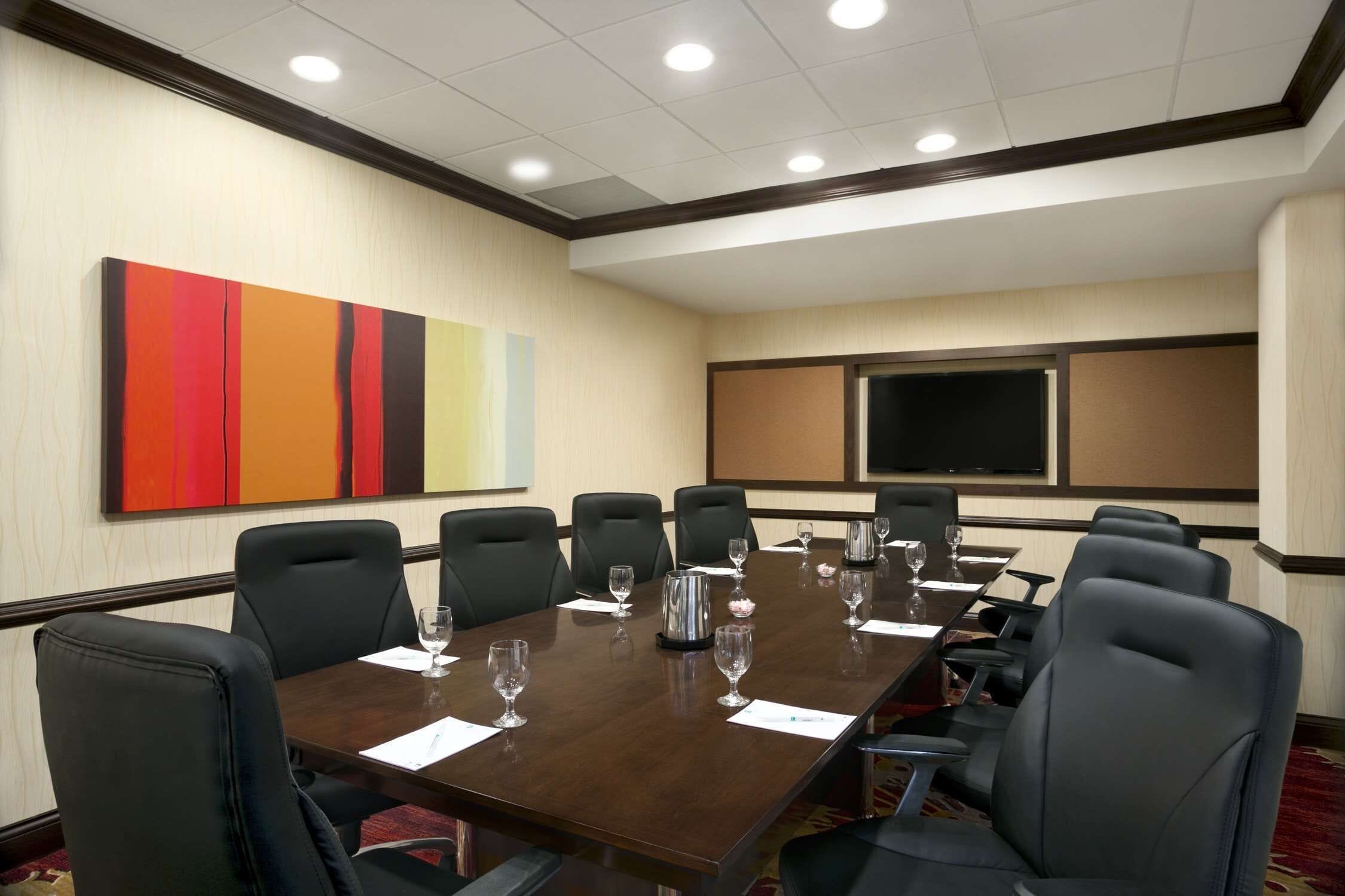 A conference room with a long table, black chairs, and a large screen on the wall.
