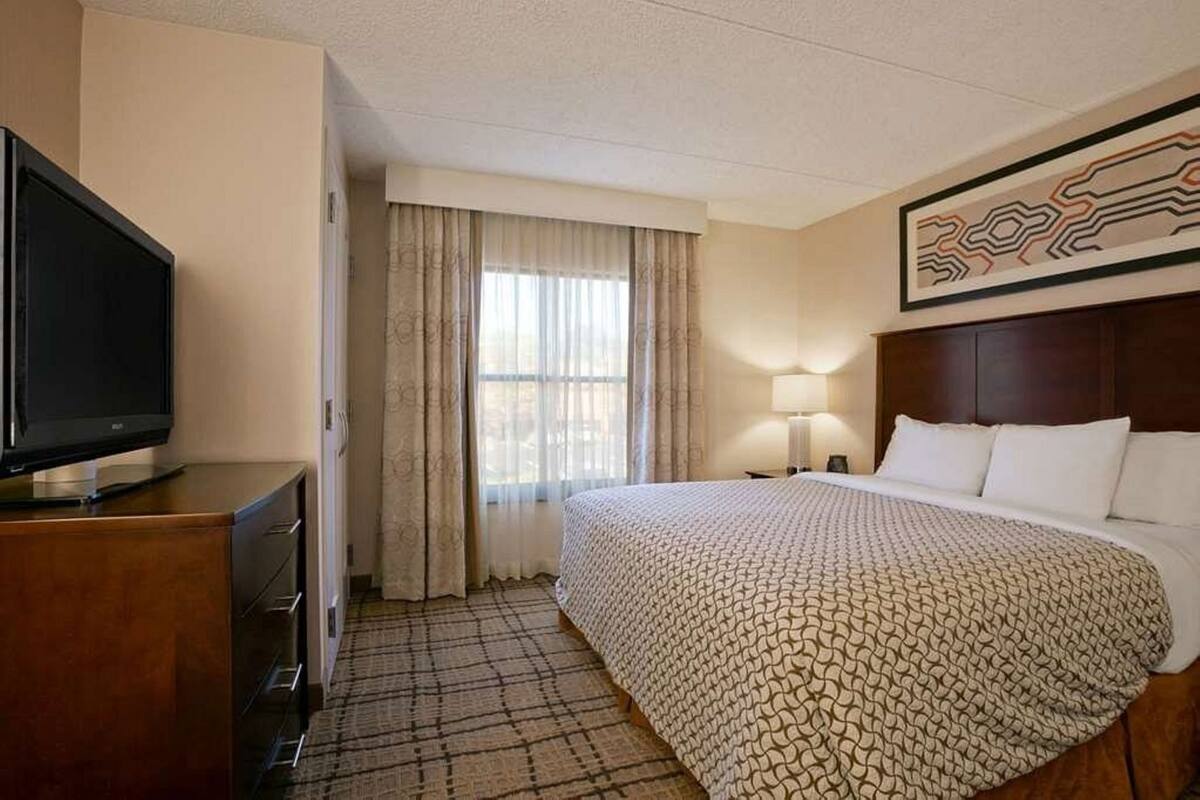 Habitación de hotel con una cama, un televisor sobre una cómoda de madera, una ventana con cortinas, una lámpara y una obra de arte en la pared.