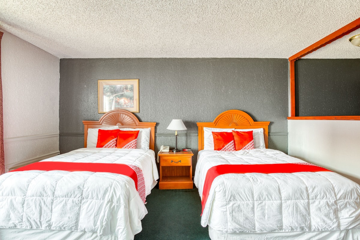 A hotel room with two beds, each with white bedding and red accents, a wooden nightstand with a lamp, and a framed picture on the wall.