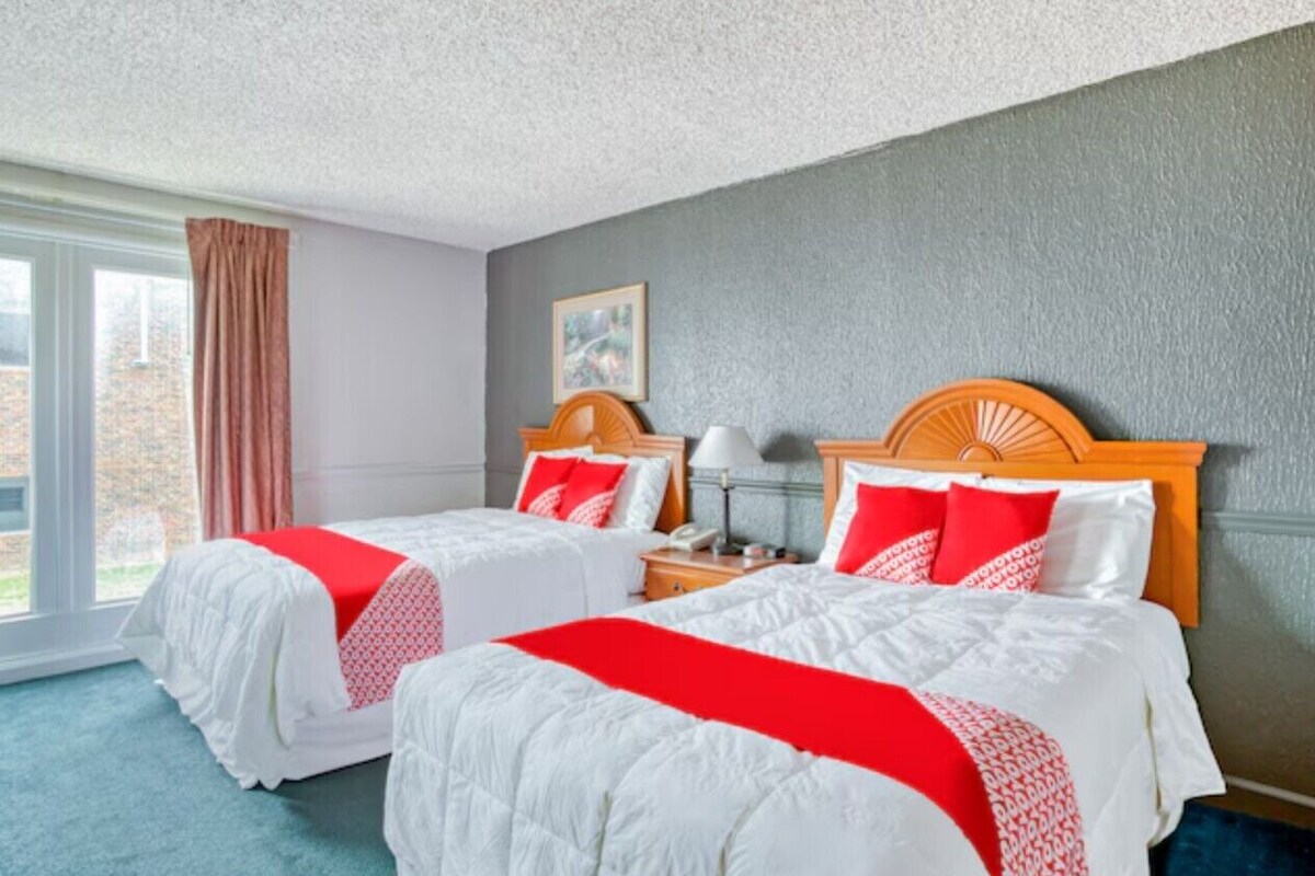 A hotel room with two beds, a window with curtains, a bedside table with a lamp, and a framed picture on the wall.