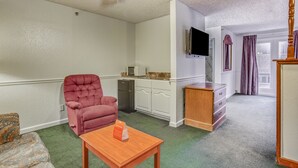 Una sala de estar con un sillón rojo, una mesa de centro de madera, una cocina con armarios blancos, un refrigerador y un televisor de pantalla plana montado en la pared.