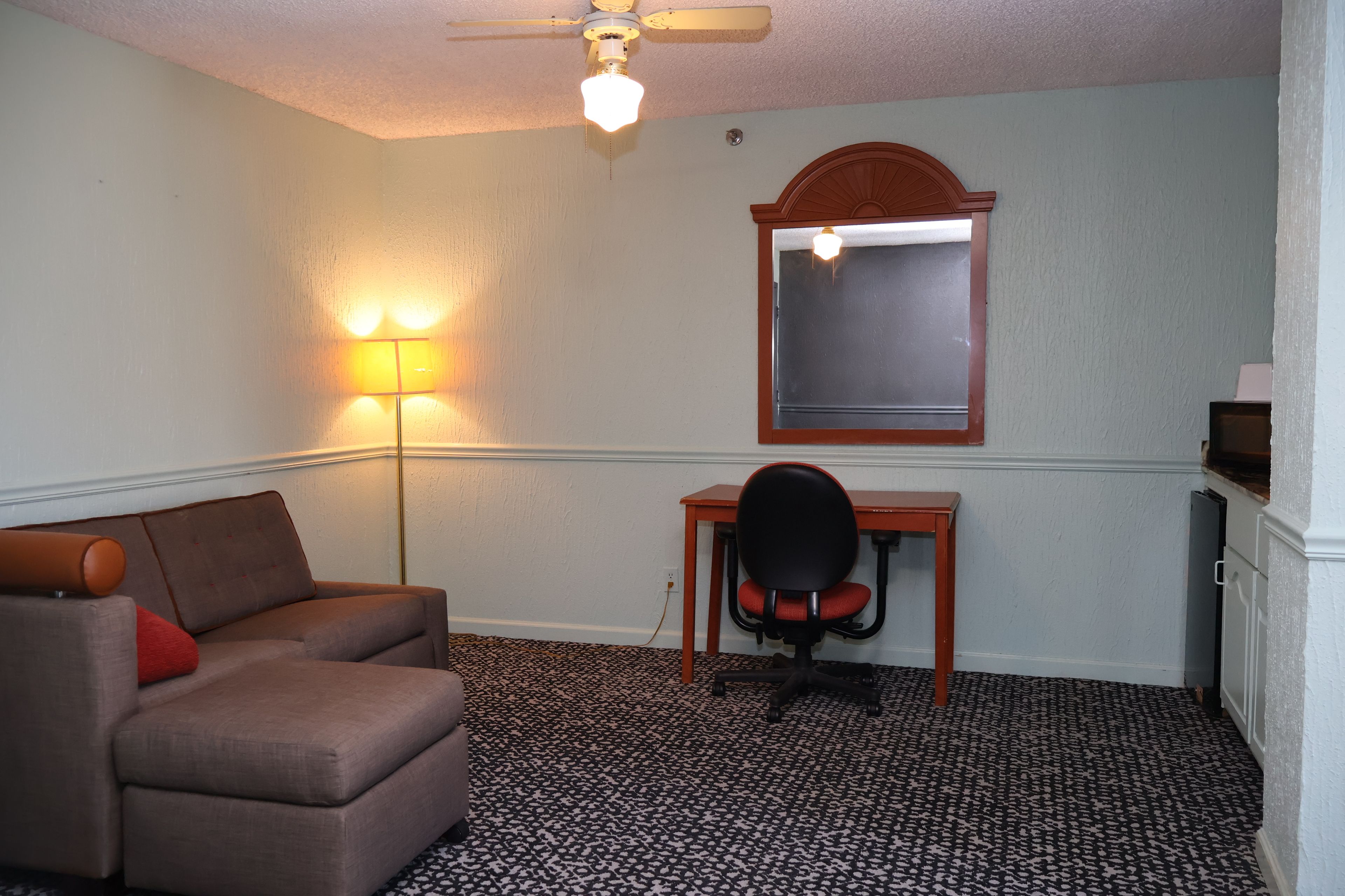 A room with a sofa, a desk, a chair, a ceiling fan, and a wall-mounted mirror.
