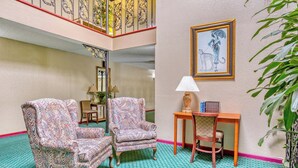 Un hall con sillones estampados, una mesita de madera con lámpara, una ilustración enmarcadas en la pared y una escalera con barandales ornamentales.