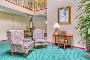 A lobby with patterned armchairs, a wooden side table with a lamp, a framed illustration on the wall, and a staircase with ornate railings. - Garden Cove Hotel Suites (Tulsa)
