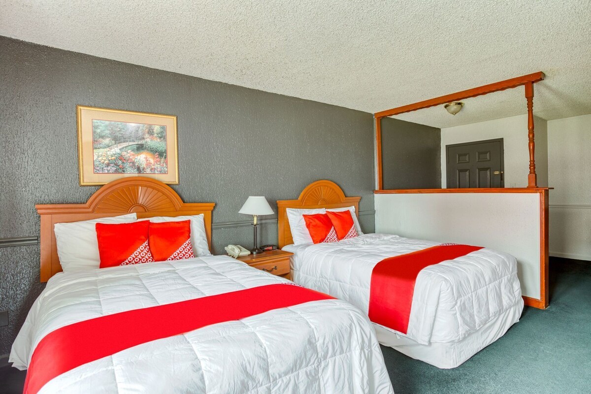 A hotel room with two beds, a large mirror, a bedside table with a lamp, and a framed picture on the wall.