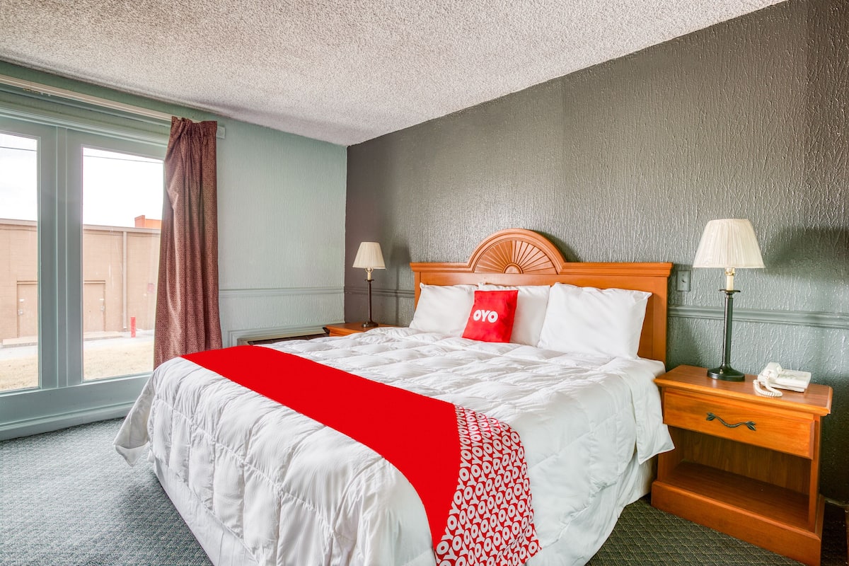 A hotel room with a large bed, a wooden headboard, two bedside tables with lamps, a red throw blanket, and a window with curtains.