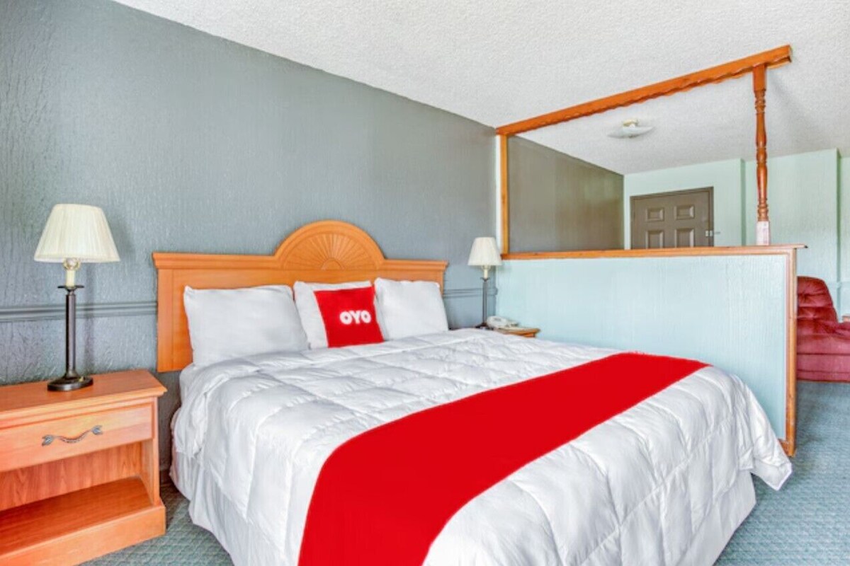 A hotel room with a bed, a nightstand, a lamp, a mirror, and a door.