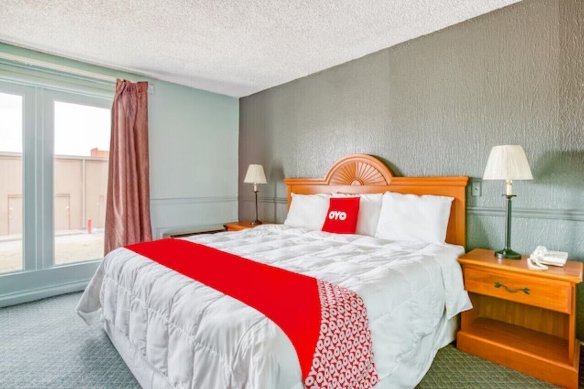 A hotel room with a bed, two bedside tables, a nightstand, a window with curtains, and a view outside.