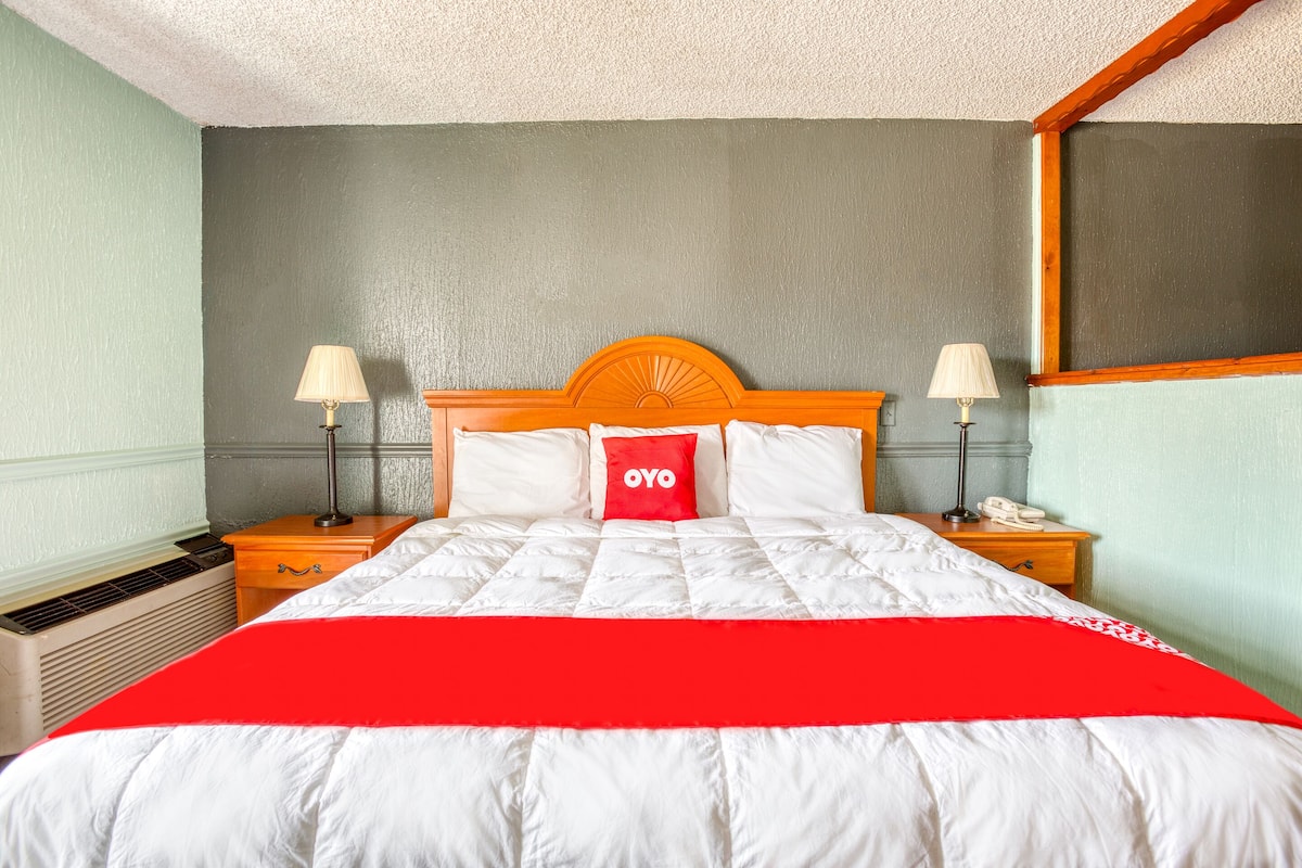 A neatly made bed with a red blanket, two bedside lamps, a wooden nightstand, and a red pillow with the word "OYO" on it.