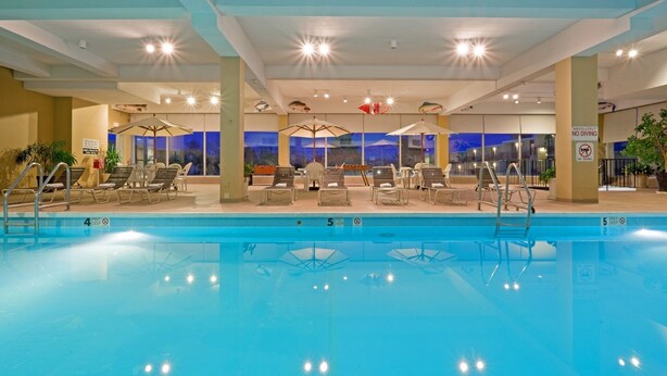 Una piscina interior con sillones de playa y sombrillas, un cartel de prohibición de buceo y vistas de la ciudad al anochecer.