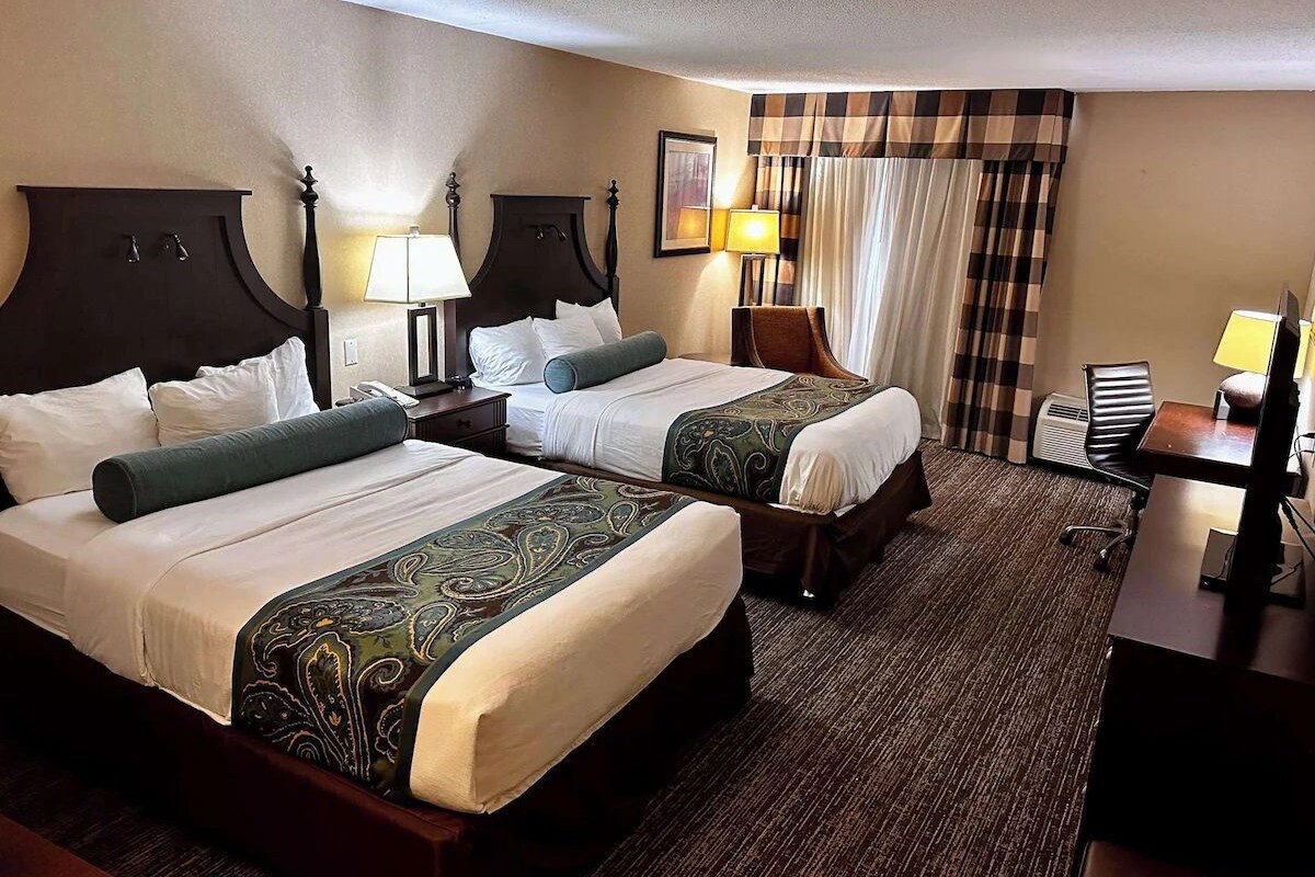 A hotel room with two beds, a desk, a chair, and a lamp.