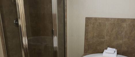 A bathroom with a glass-enclosed shower, a white bathtub, and beige tiled flooring.