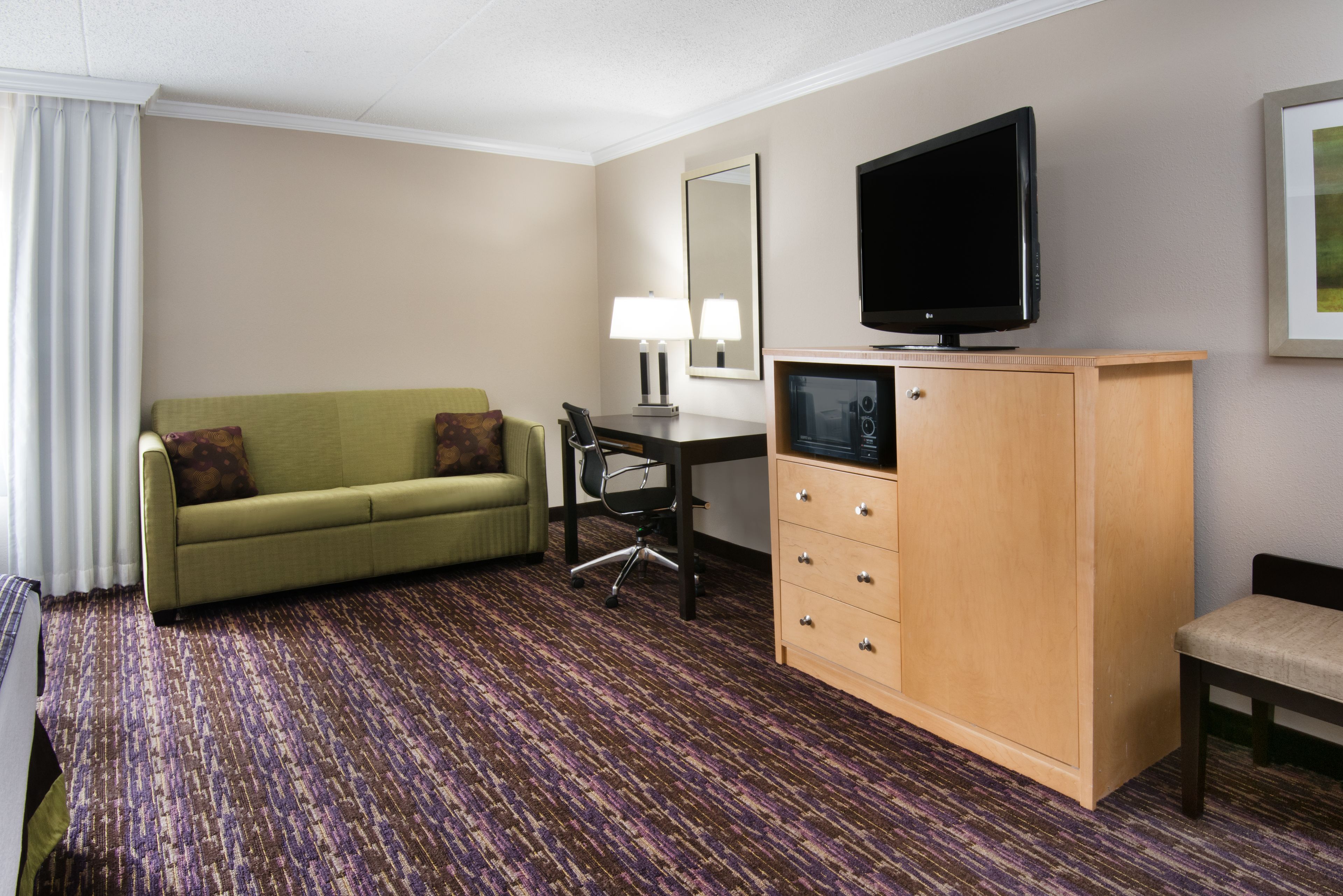 Habitación de hotel con un sofá verde, un aparador de madera con cajones, un escritorio con una silla, un televisor y un espejo grande en la pared.