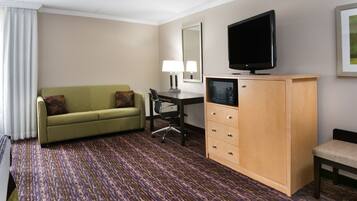 Habitación de hotel con un sofá verde, un aparador de madera con cajones, un escritorio con una silla, un televisor y un espejo grande en la pared.