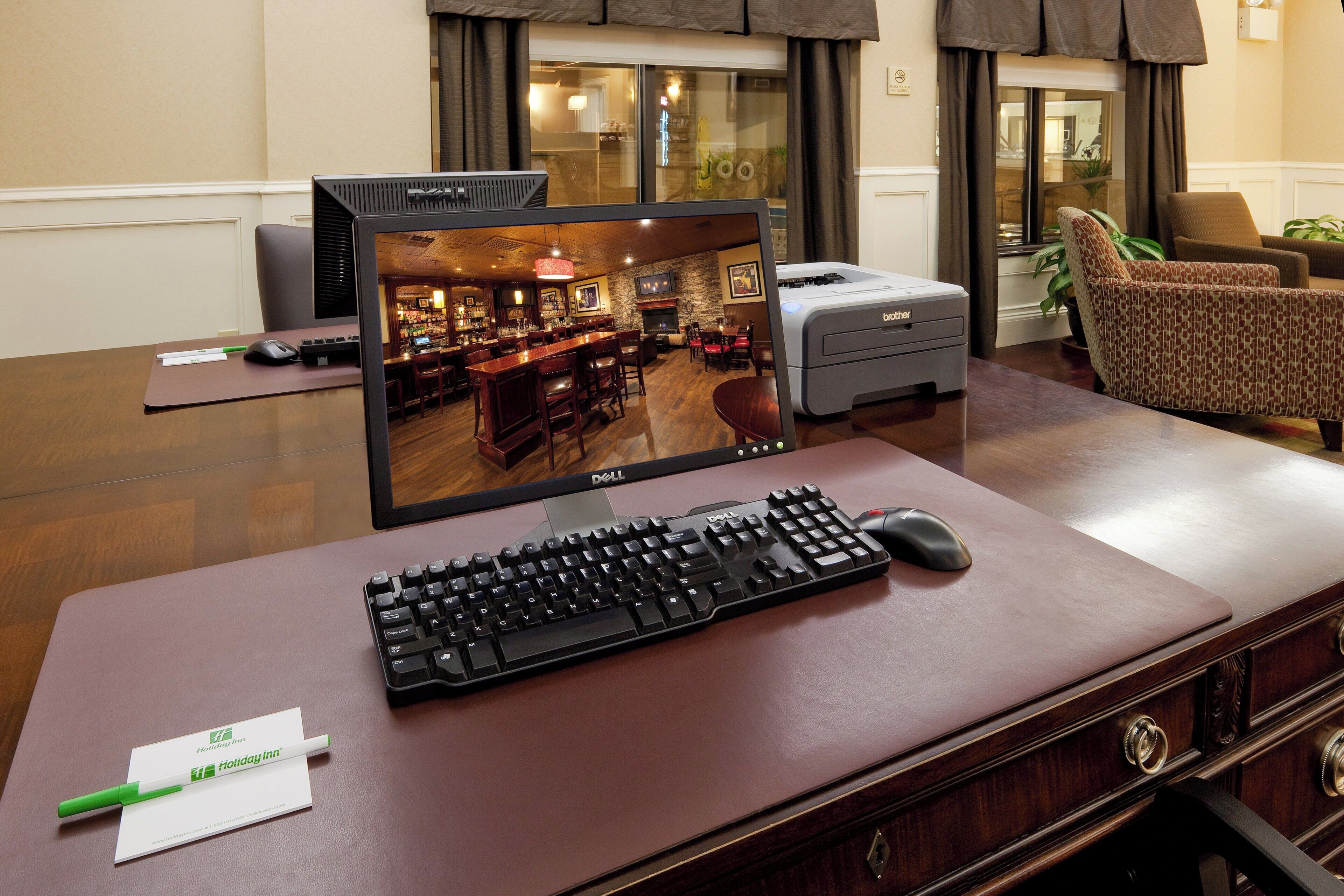 A hotel room with a desk, a computer monitor, a keyboard, a mouse, a printer, and a chair.