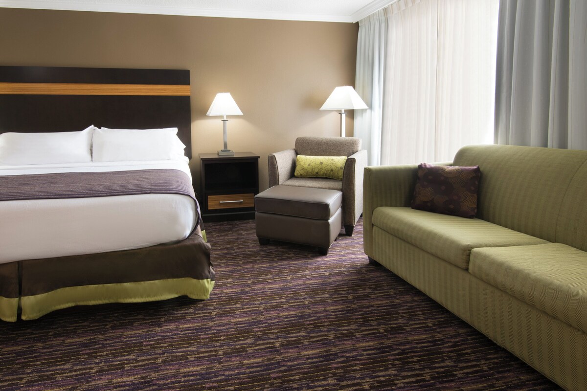 A hotel room with a bed, a chair, a sofa, a nightstand, and a lamp.
