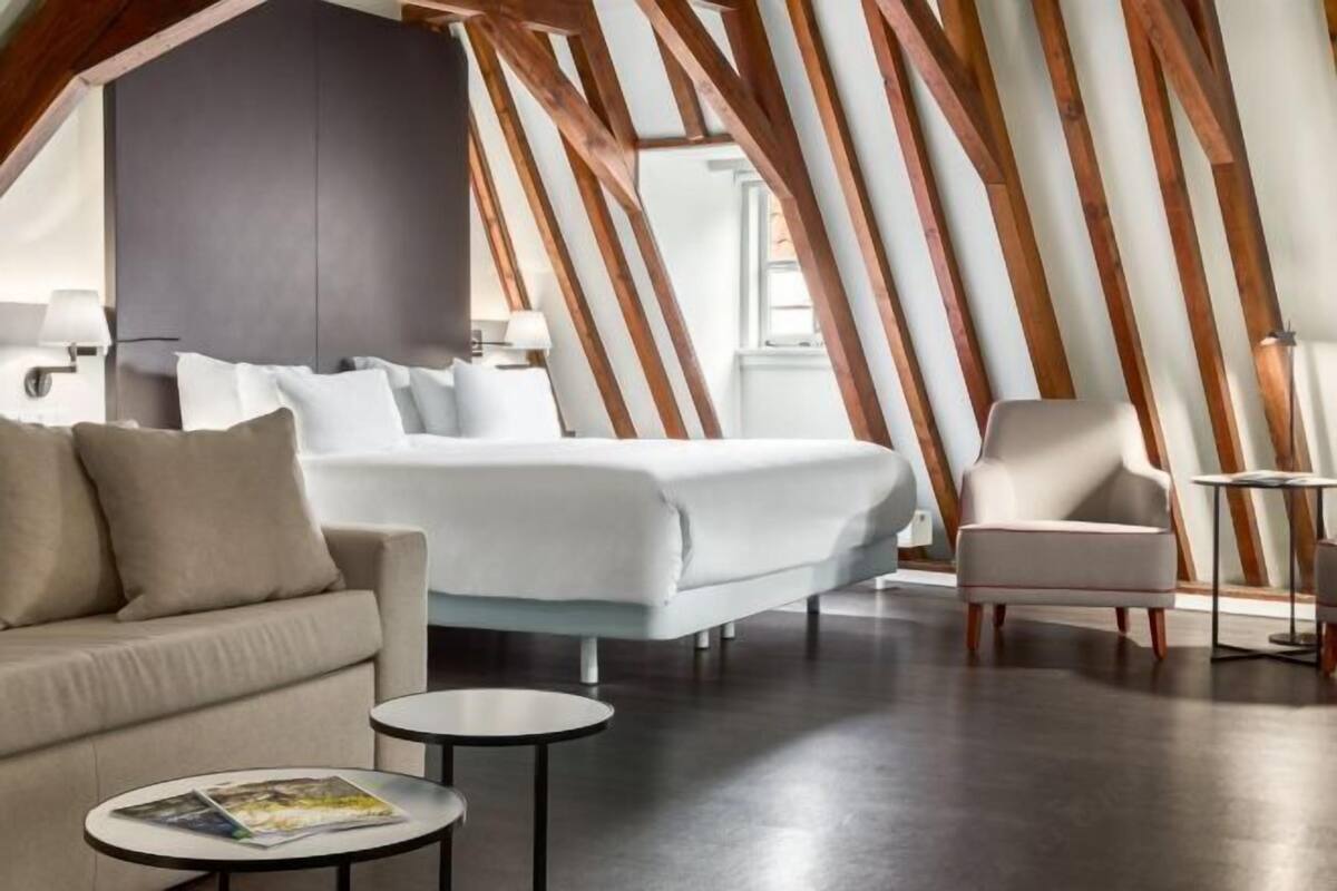 A modern hotel room with a bed, a sofa, a chair, a small table, and a large window.
