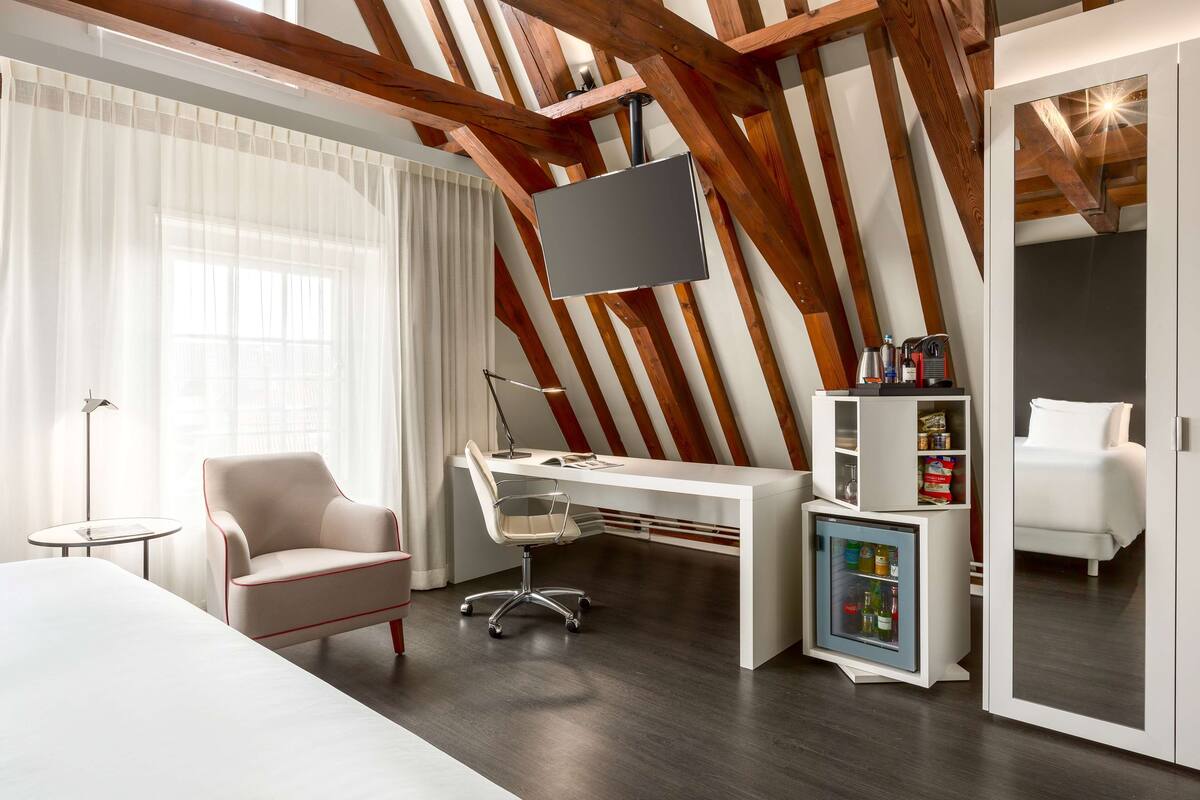 A modern hotel room with a wooden ceiling, a large bed, a desk with a chair, a small refrigerator, and a bedside table.