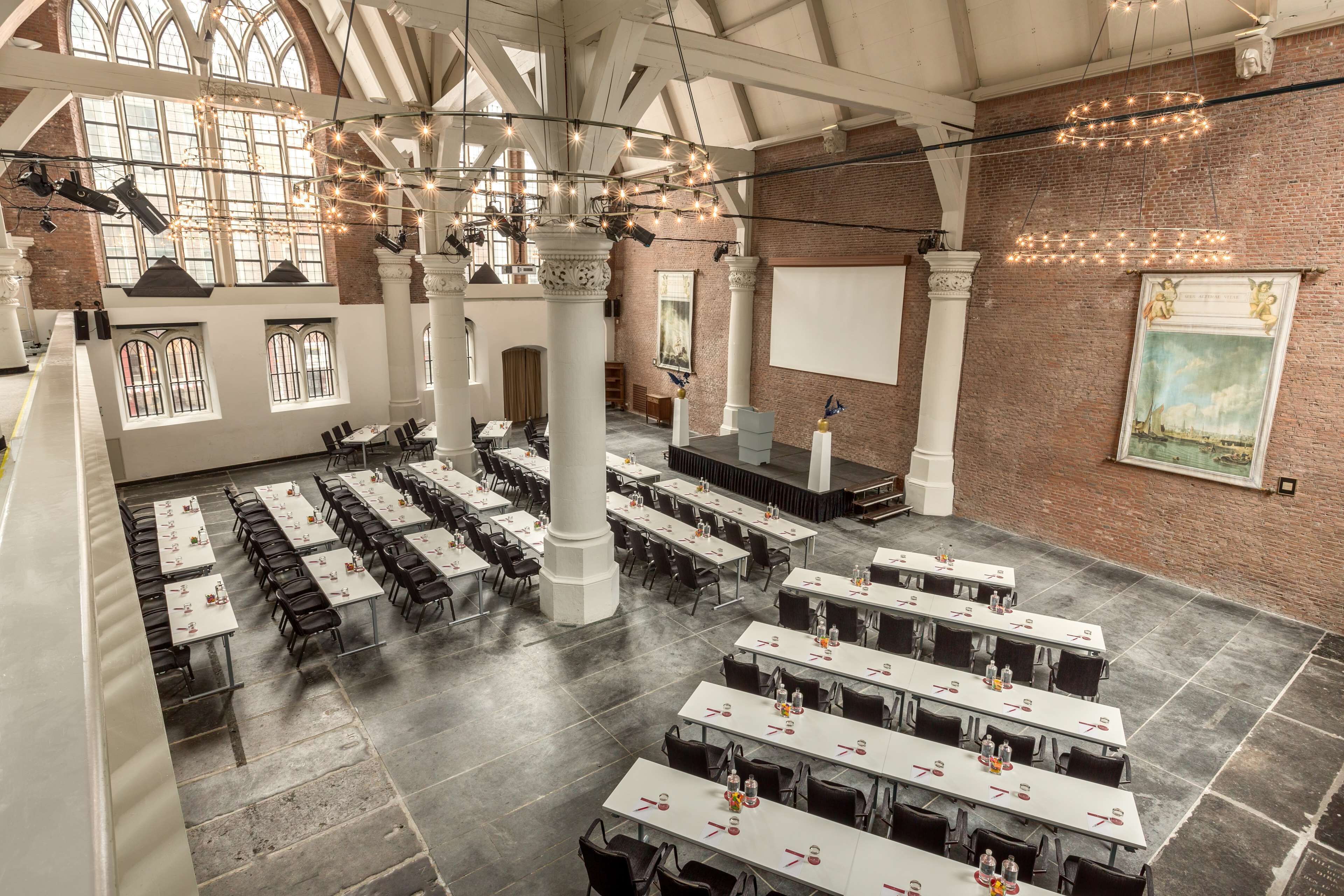 A spacious hall with high ceilings, large windows, and a brick wall. Tables are arranged in rows, and there are columns supporting the ceiling.