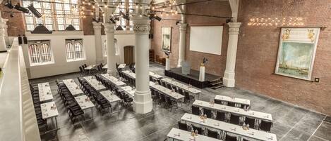 A spacious hall with high ceilings, large windows, and a brick wall. Tables are arranged in rows, and there are columns supporting the ceiling.