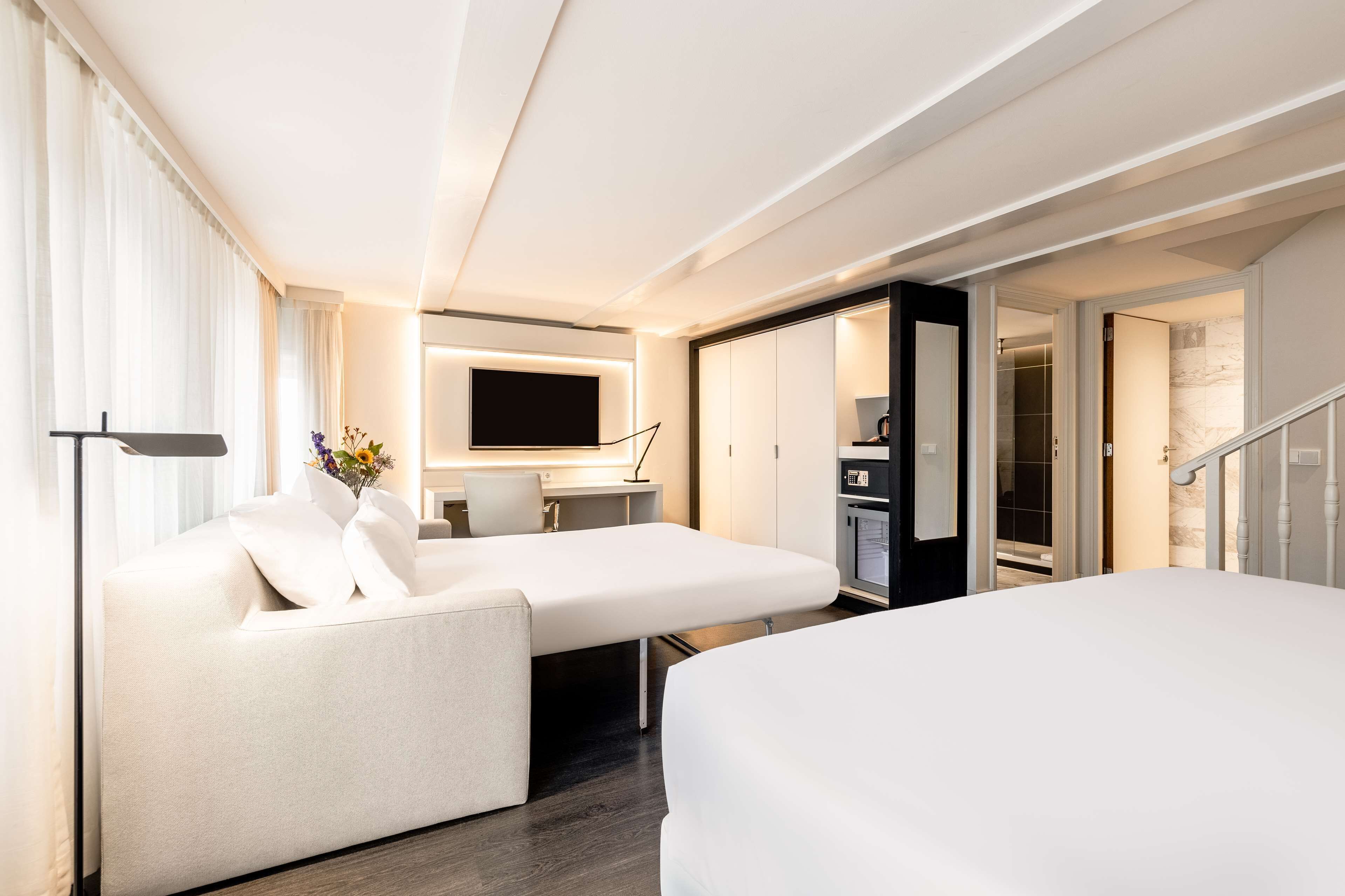 Ein modernes Hotelzimmer mit einer Couch, einem Bett, einem Schreibtisch und einem Flachbildfernseher sowie einem begehbaren Kleiderschrank.