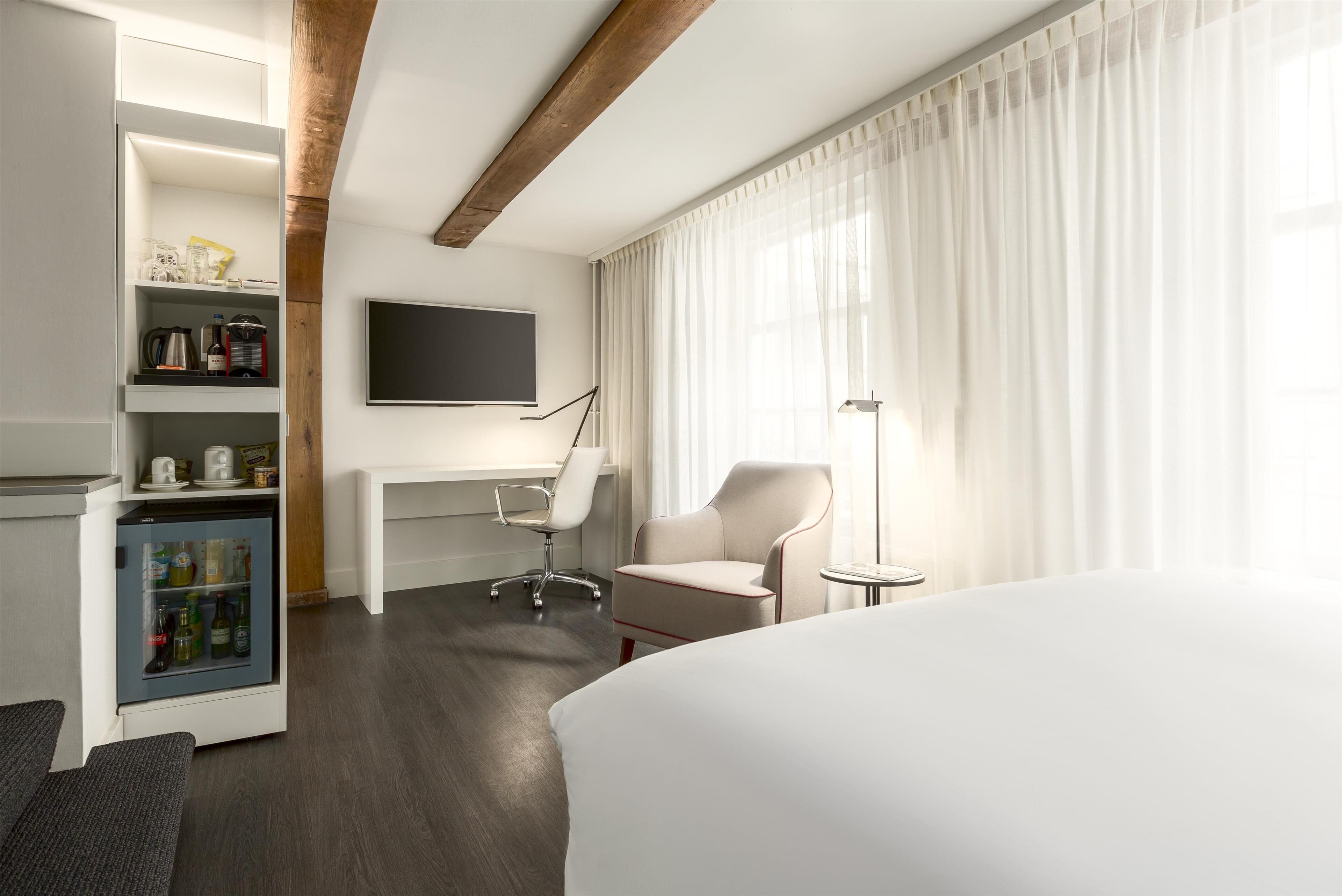 A modern hotel room with a bed, a desk, a chair, a small refrigerator, and a TV on the wall.