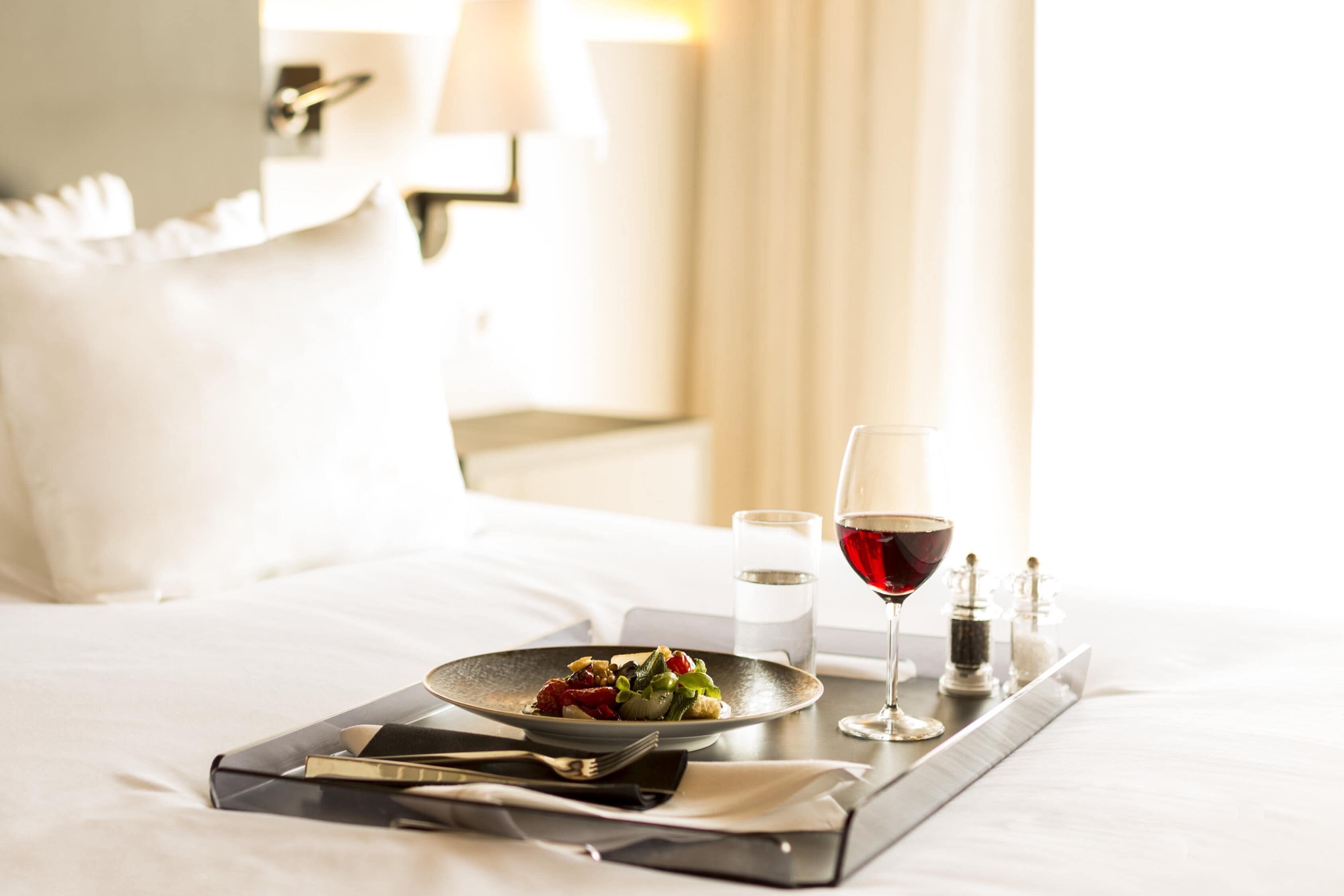 A neatly made bed with a tray containing a plate of food, a glass of red wine, and a water glass.