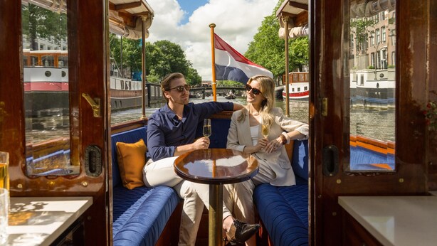 A couple seated at a table inside a boat with a view of the river and buildings. - NH Collection Amsterdam Barbizon Palace (Amsterdam)