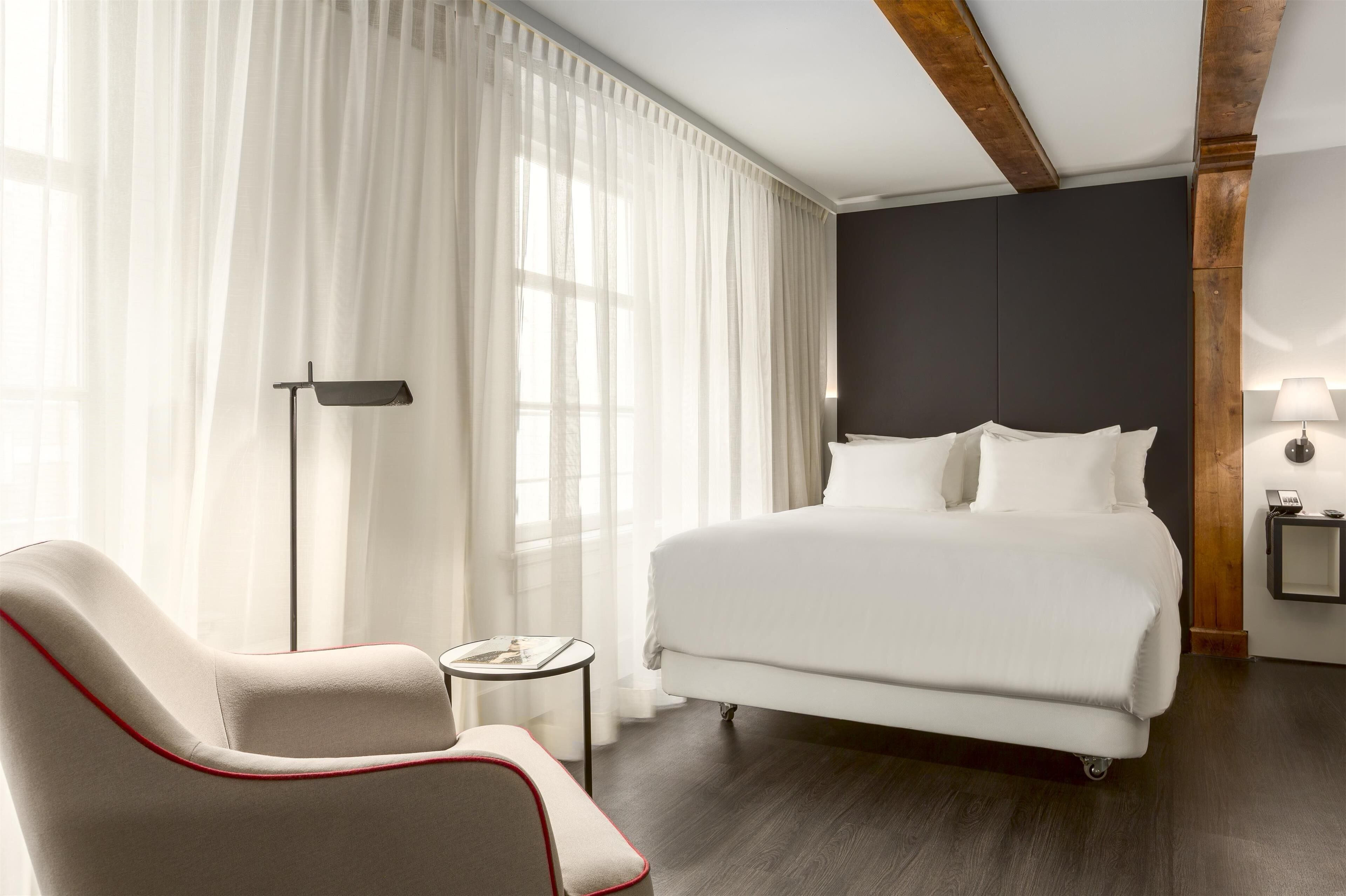A modern hotel room with a large bed, a chair, a bedside table, and a lamp.