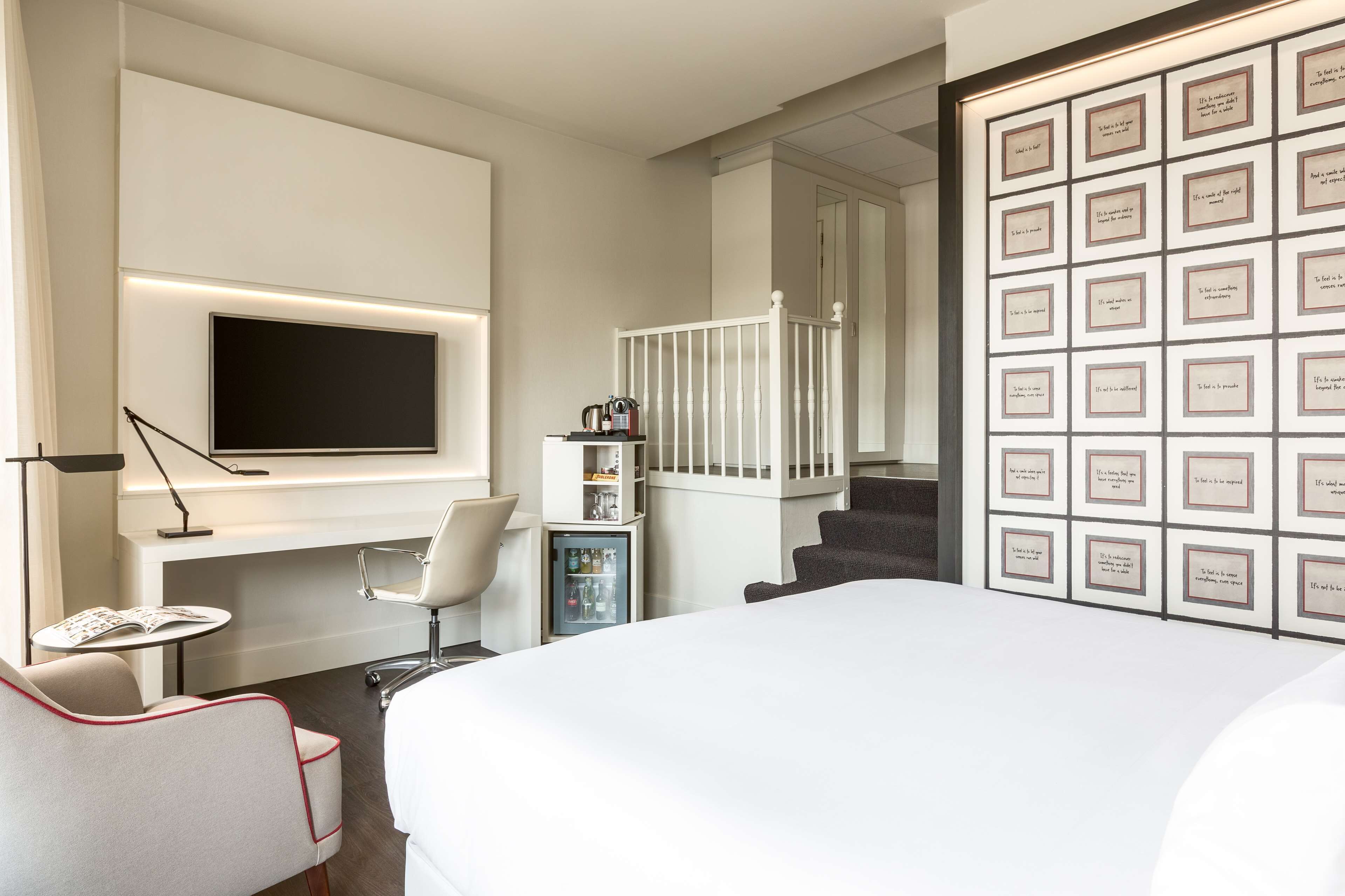 A modern hotel room with a large bed, a flat-screen TV mounted on the wall, a small table with a chair, a minibar, and a staircase leading to another room.