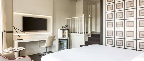 A modern hotel room with a large bed, a flat-screen TV mounted on the wall, a small table with a chair, a minibar, and a staircase leading to another room.