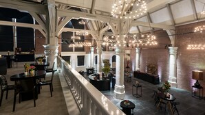A spacious hotel lobby with high ceilings, exposed brick walls, and a series of columns. There are multiple seating areas with tables and chairs, and decorative lighting fixtures. - NH Collection Amsterdam Barbizon Palace (Amsterdam)
