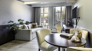A modern hotel room with a sofa, dining table, and a large window offering a city view. - NH Collection Amsterdam Barbizon Palace (Amsterdam)