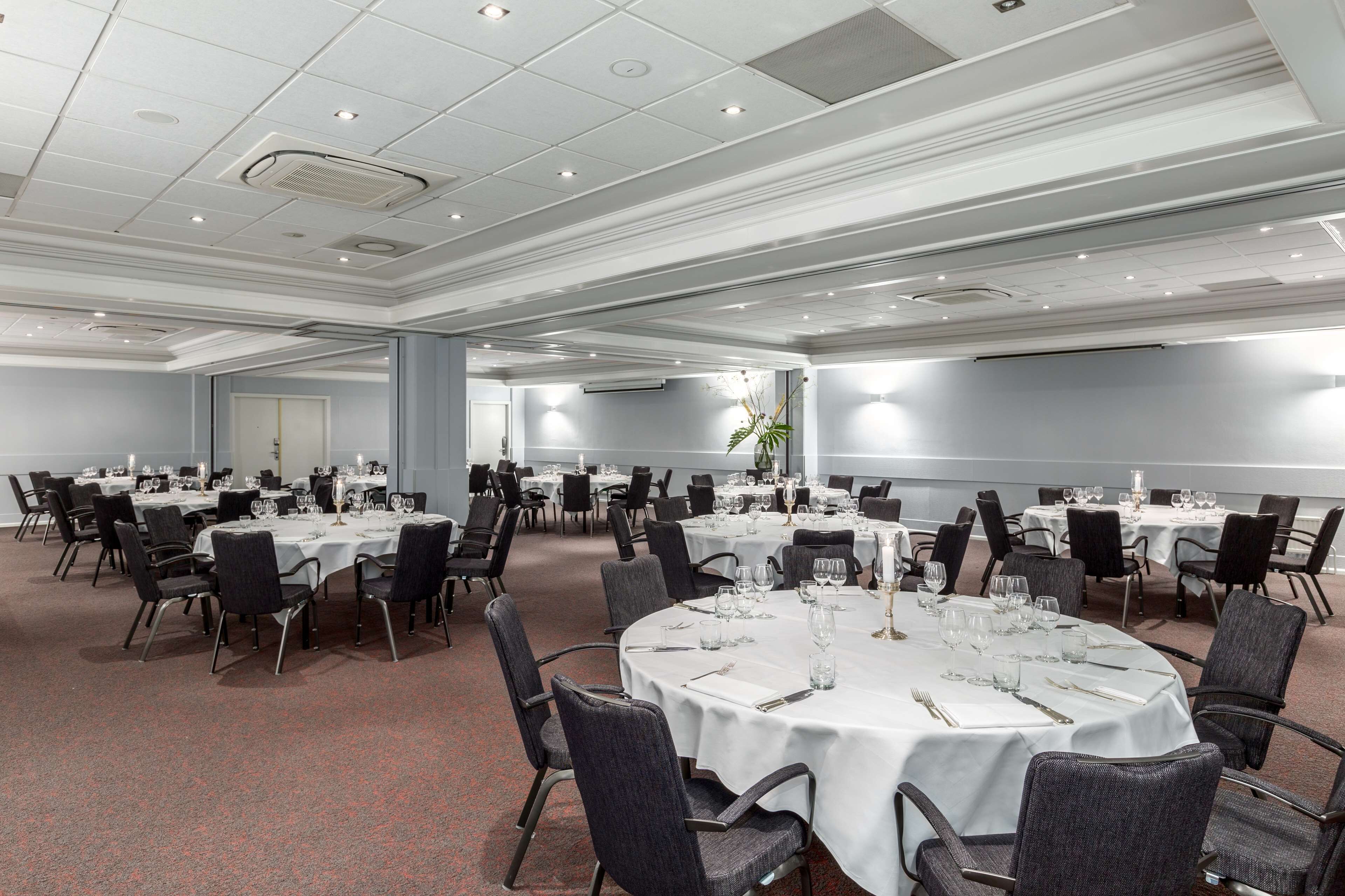 Une grande salle de conférence dotée de nombreuses tables rondes, chacune recouverte d’une nappe blanche et agrémentée de chaises et de couverts.