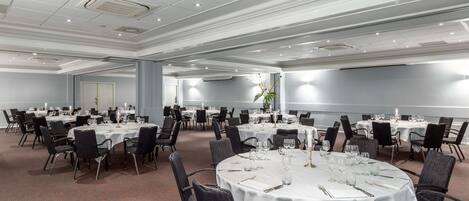 Une grande salle de conférence dotée de nombreuses tables rondes, chacune recouverte d’une nappe blanche et agrémentée de chaises et de couverts.
