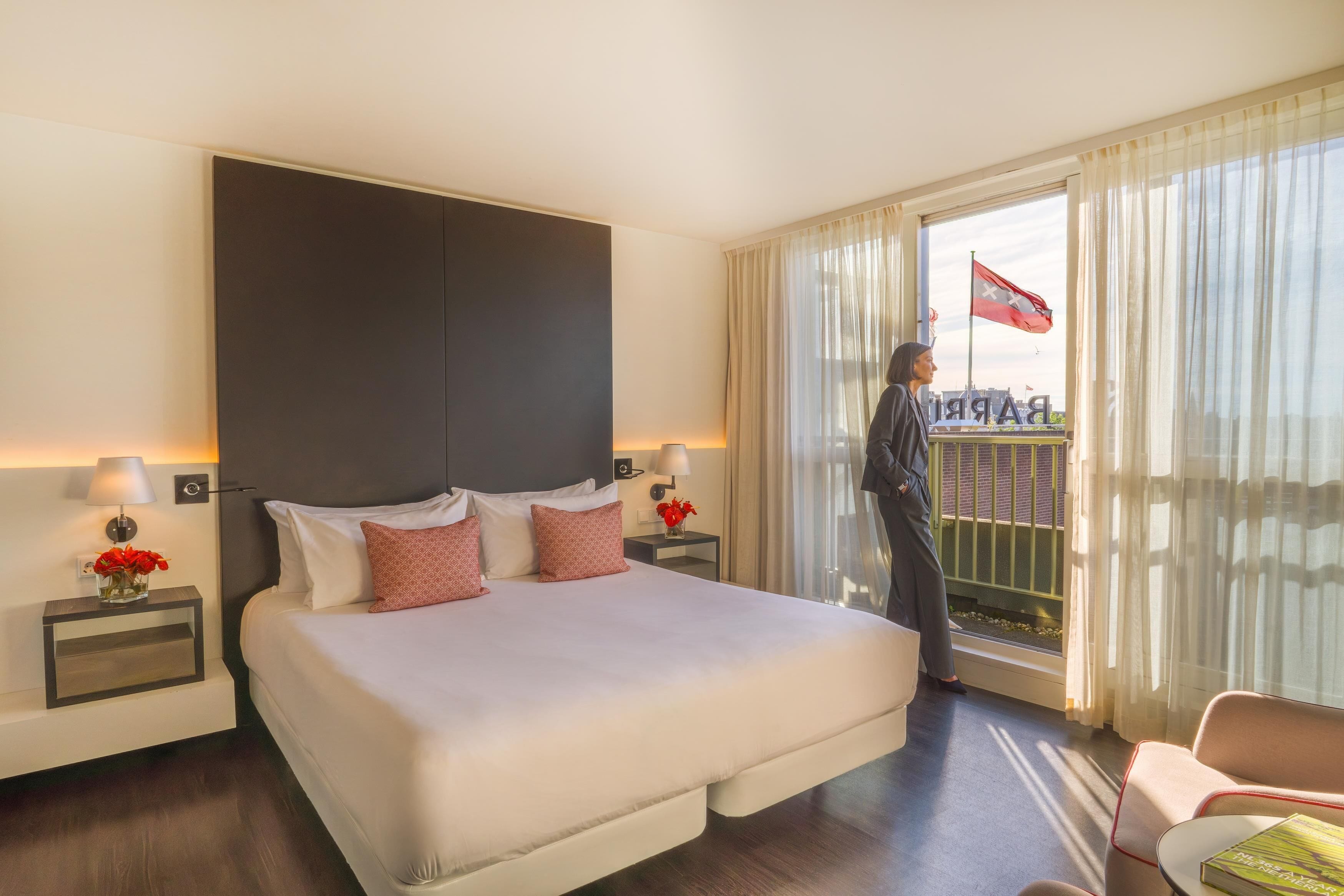 Habitación de hotel con una cama grande, dos mesitas de noche con lámparas, una mesita de noche con un jarrón de flores y un balcón con una bandera.