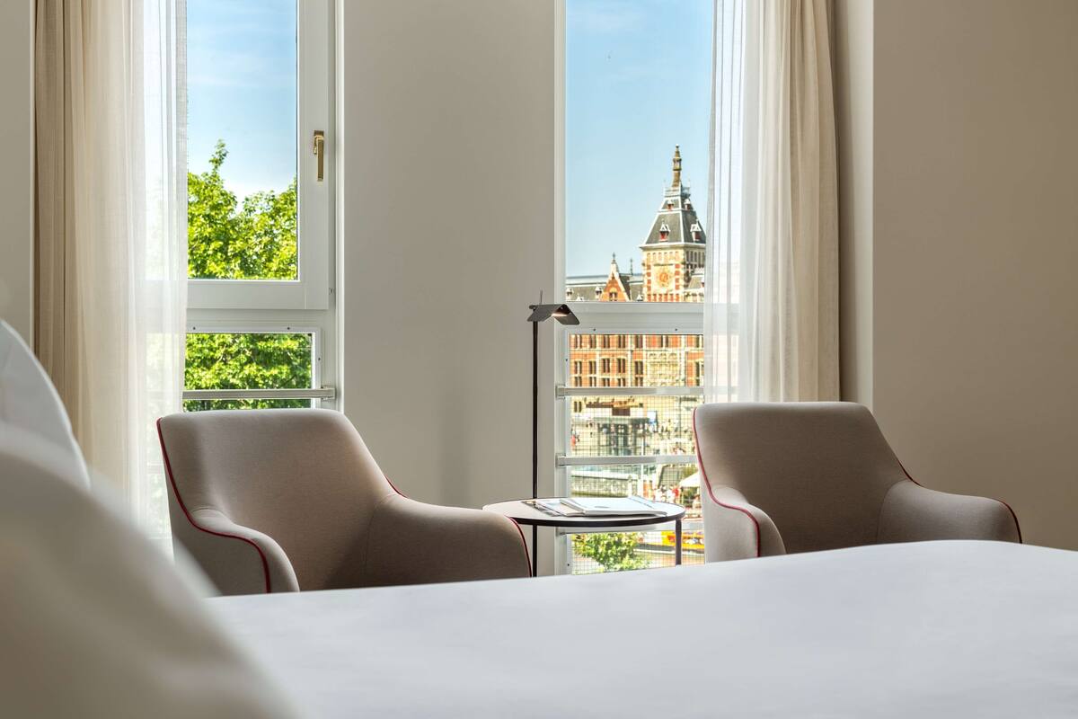 A hotel room with two chairs, a small table, and a view of a cityscape through large windows.