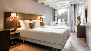 A modern hotel room with a large bed, bedside lamps, a desk, and a view of the city through large windows. - NH Collection Amsterdam Barbizon Palace (Amsterdam)