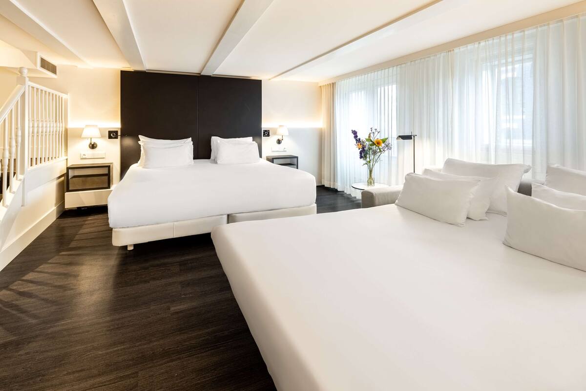 A modern hotel room with a large bed, a smaller bed, a nightstand, a lamp, a vase with flowers, and a view of the outdoors through sheer curtains.
