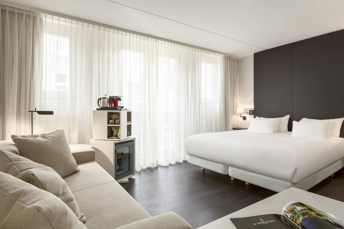 A modern hotel room with a large bed, a sofa, a small side table, and a bookshelf.
