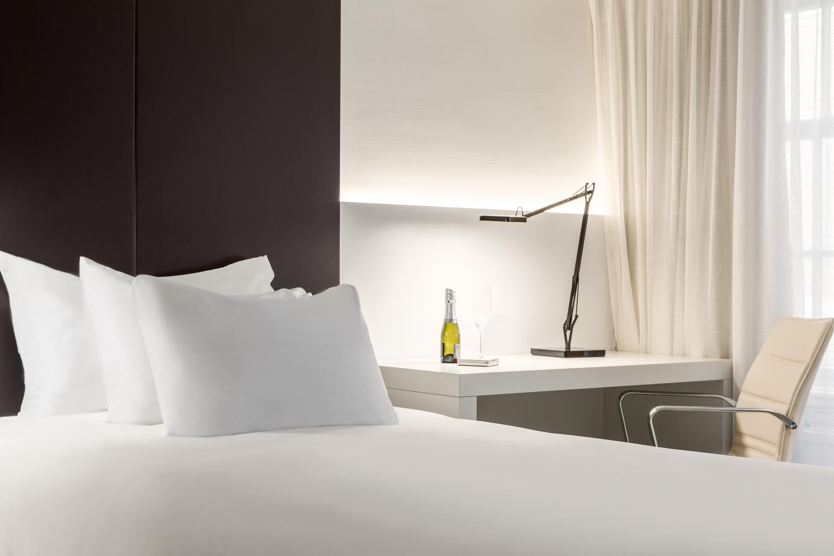 Habitación de hotel moderna con una cama grande, una mesita de noche con una botella y un vaso, una lámpara de escritorio y una silla.