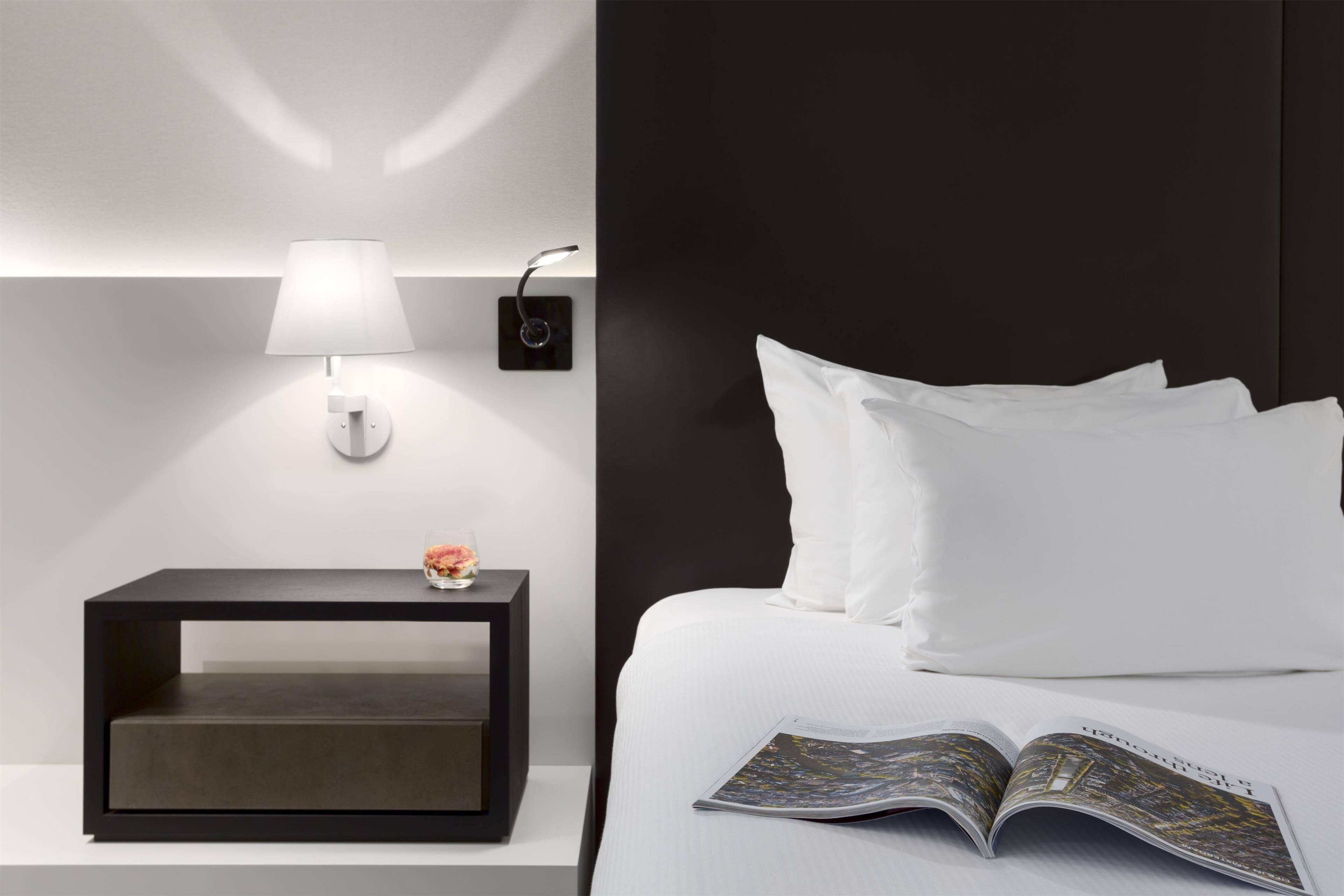 A modern hotel room with a bed, a bedside table with a lamp, and a magazine on the bed.